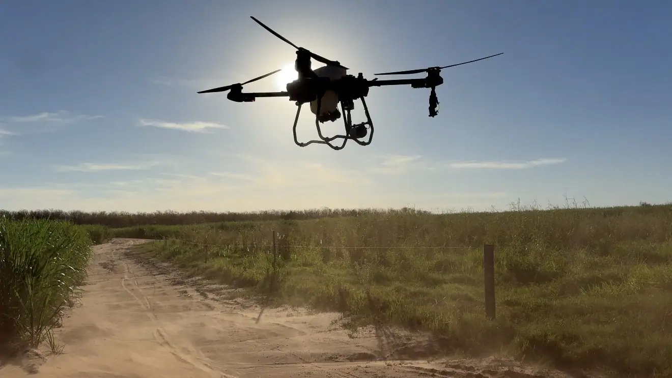 Drone agrícola em voo sobre uma estrada de terra ao lado da lavoura, com o sol atrás do equipamento