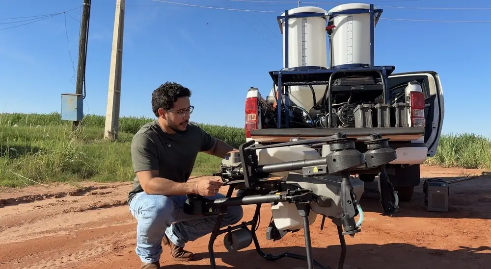 Lucas Fernando Peres agachado conferindo o drone agrícola em frente à camionete com a estrutura móvel de abastecimento