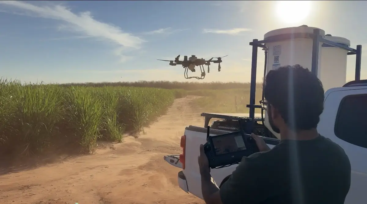 Lucas Fernando Peres de costas segurando o rádio controle enquanto o drone agrícola sobrevoa a estrada ao lado da lavoura de cana
