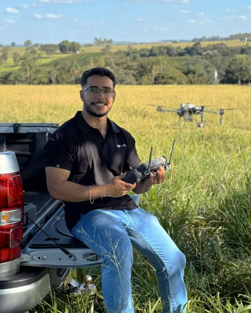 Lucas Fernando Peres segurando o rádio controle, com um drone agrícola em voo ao fundo na lavoura