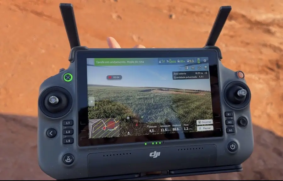 Rádio controle de drone agrícola em mãos, com a tela exibindo a tarefa de pulverização em andamento, área coberta e parâmetros de voo